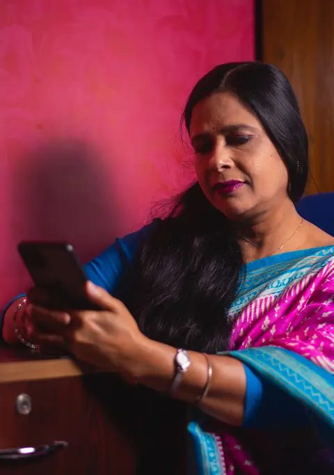 An Indian woman in a colourful saree sits indoors checking her smartphone with a focused expression.