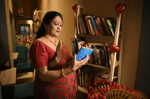 A South Asian woman in a red saree stands by a bookshelf at home, absorbed in reading a paperback.