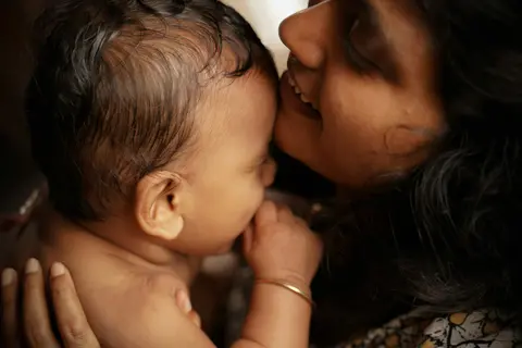 A South Asian mother tenderly kisses her baby's cheek in close-up, warm golden light and a plain dark background.