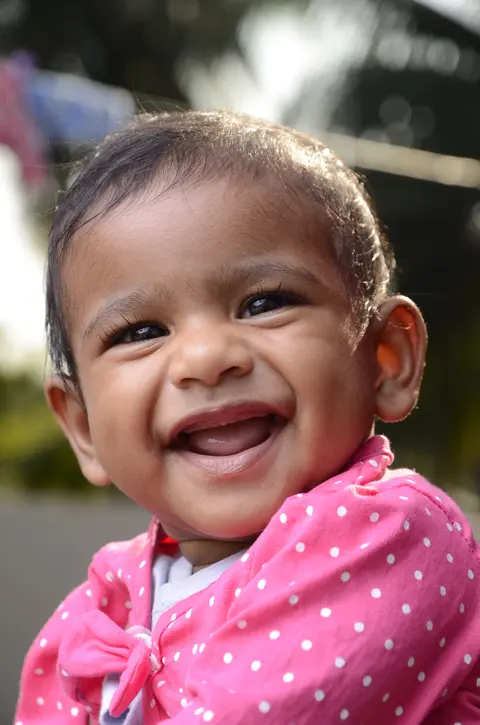 Close-up of a South Asian baby girl in a pink polka-dot outfit laughing joyfully outdoors in soft natural light.