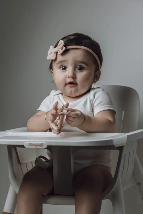 A baby girl in a bow headband sits upright in a white high chair, holding a piece of food up to her mouth with both hands.