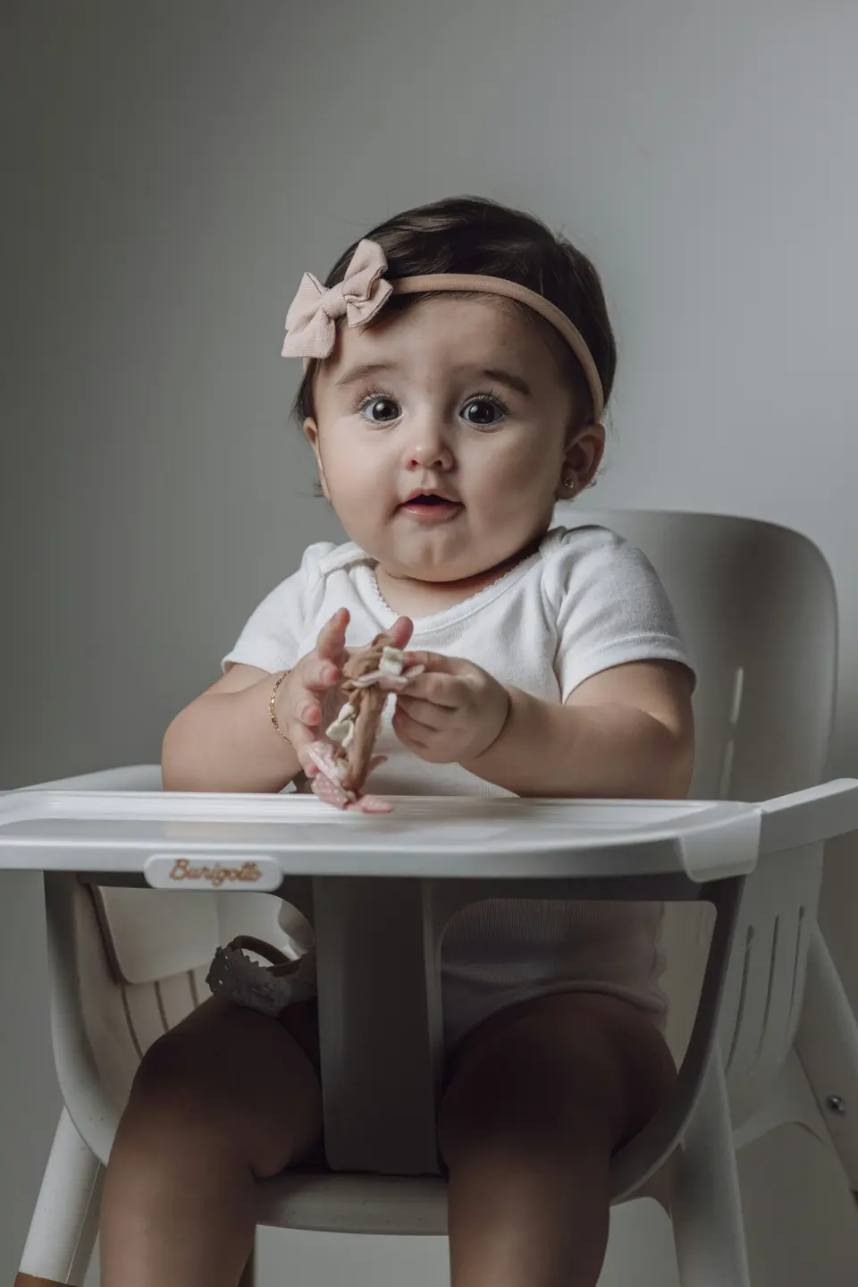 A baby girl in a bow headband sits upright in a white high chair, holding a piece of food up to her mouth with both hands.