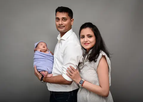 An Indian couple stand back-to-back for a studio portrait, the father securely cradling their swaddled newborn in a grey wrap and cap while the mother smiles at the camera.