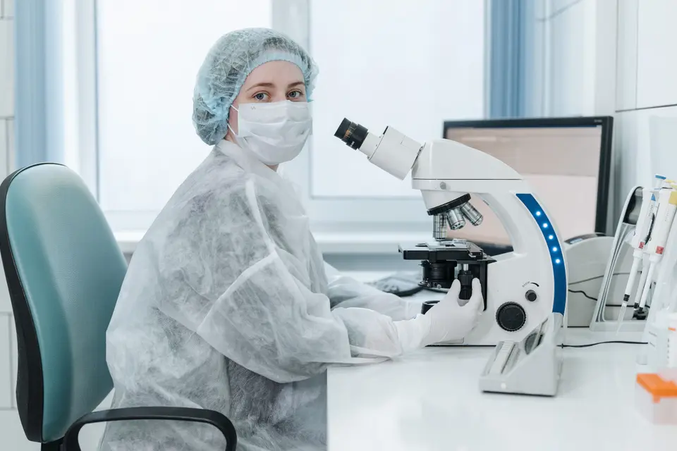A masked, gowned lab technician sits beside a trinocular microscope in a medical laboratory.