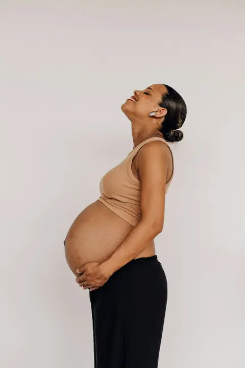 A pregnant Black woman in a tan tank top stands in profile against a grey studio backdrop, head tilted back and hand on her belly