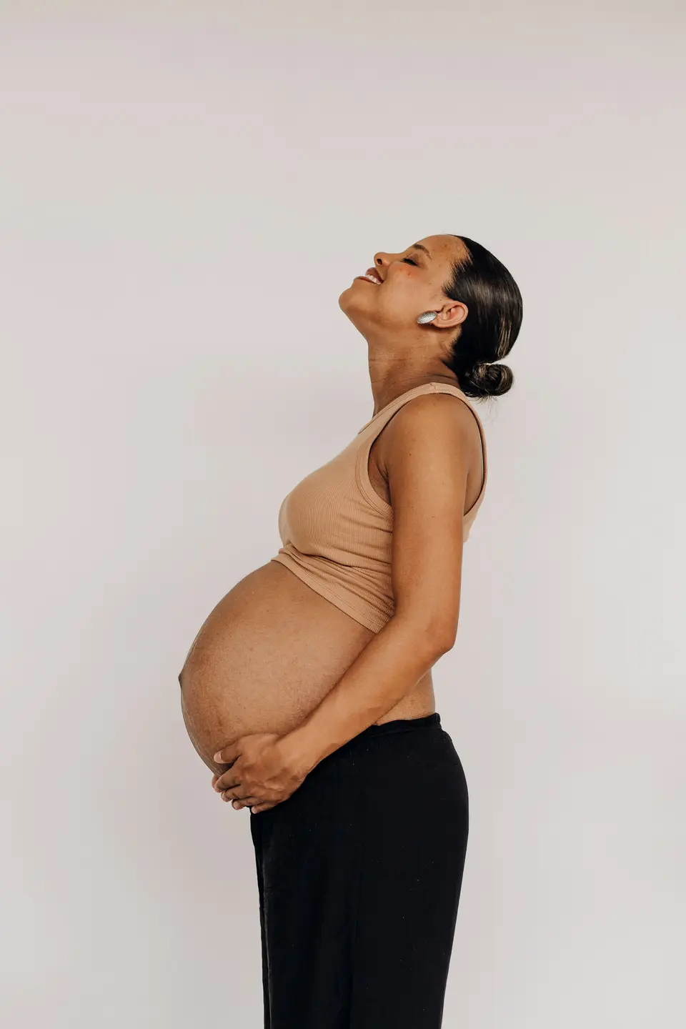 A pregnant Black woman in a tan tank top stands in profile against a grey studio backdrop, head tilted back and hand on her belly
