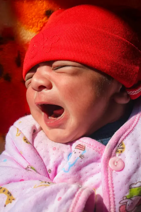 A South Asian baby in a red knit cap yawns widely with eyes scrunched shut, held by a caregiver in warm sunlight.