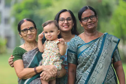 Four generations of Indian women, from grandmother to granddaughter, stand together smiling in a garden.
