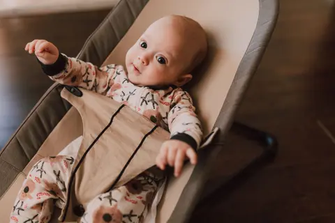 A baby lies awake and alert in a stationary bouncer seat, arms raised, wearing a floral onesie with no jewellery, face clearly toward the camera.