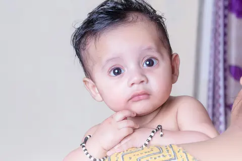 Close-up portrait of an Indian baby's face and dark hair, wide curious eyes looking at the camera, wearing a black-and-white beaded wrist bracelet.