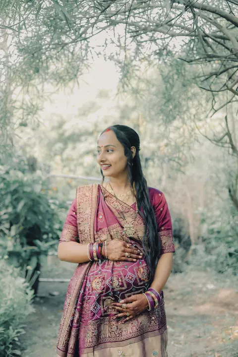 A smiling pregnant woman in a maroon sari with gold trim stands among bare trees, one hand resting on her belly