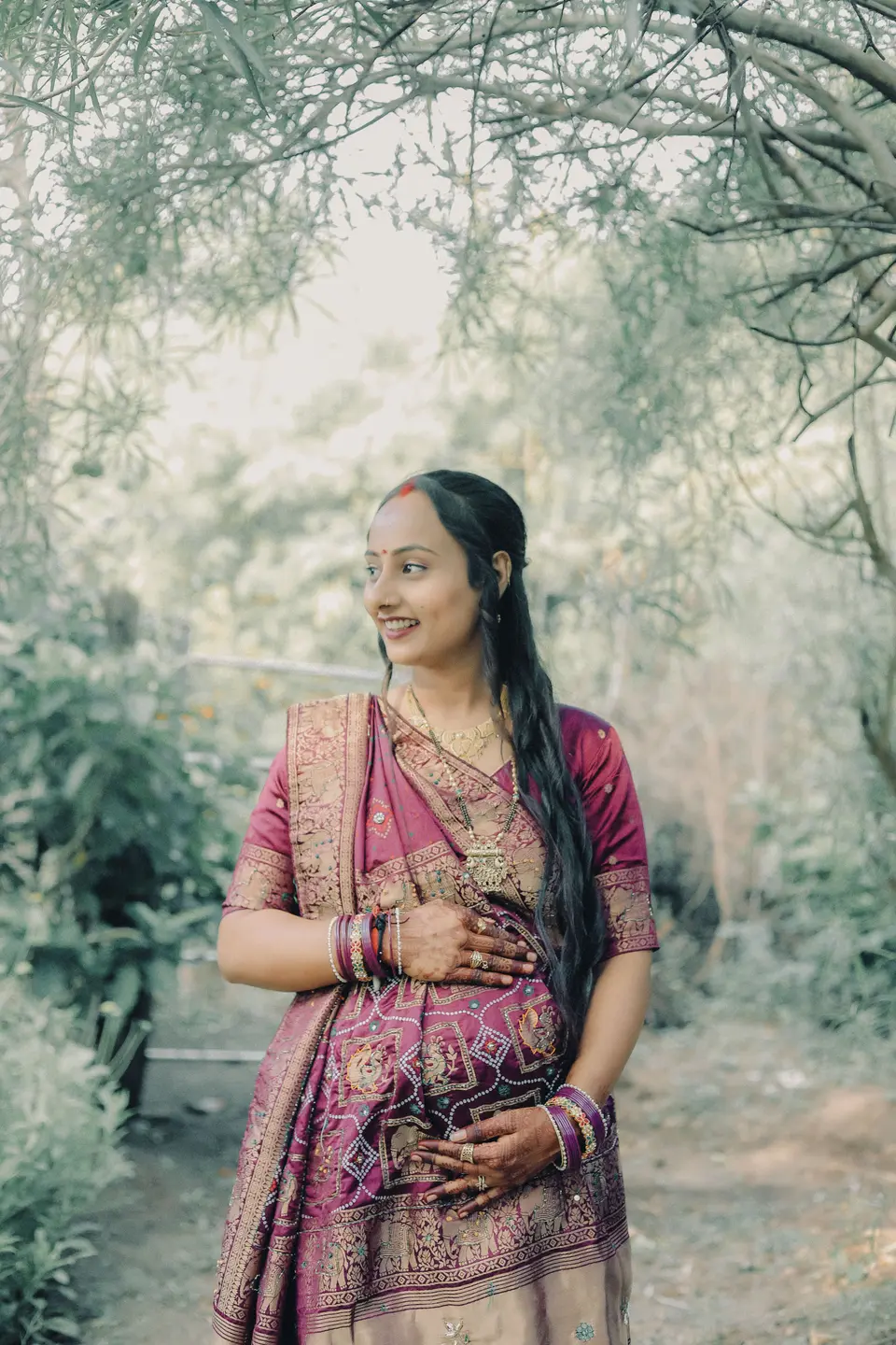 A smiling pregnant woman in a maroon sari with gold trim stands among bare trees, one hand resting on her belly