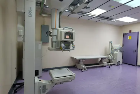 An empty hospital imaging room with an X-ray/fluoroscopy machine positioned over an examination table.