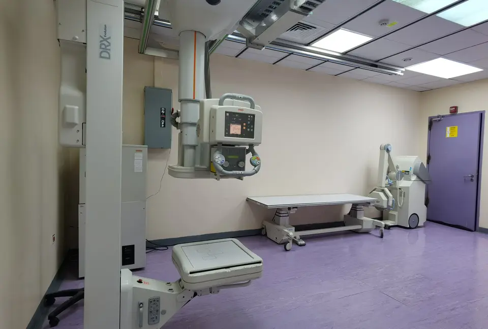 An empty hospital imaging room with an X-ray/fluoroscopy machine positioned over an examination table.