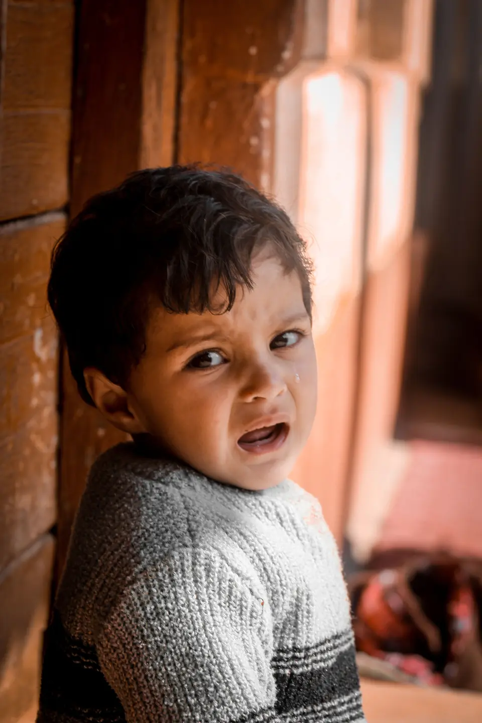 A South Asian toddler boy in a gray knitted sweater cries with a tear on his cheek, glancing back near a wooden doorway.