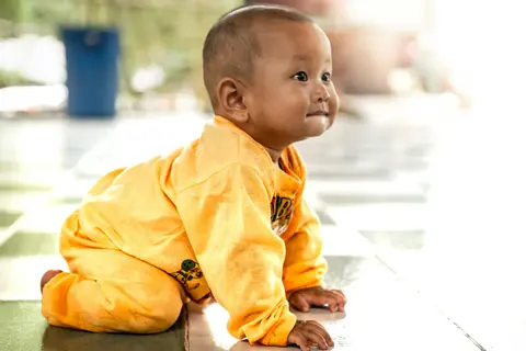 A baby in a yellow onesie crawls on hands and knees across a tiled floor, looking up and smiling.