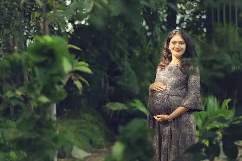 A smiling pregnant woman in a shimmering grey dress stands in a leafy garden with one hand resting on her belly