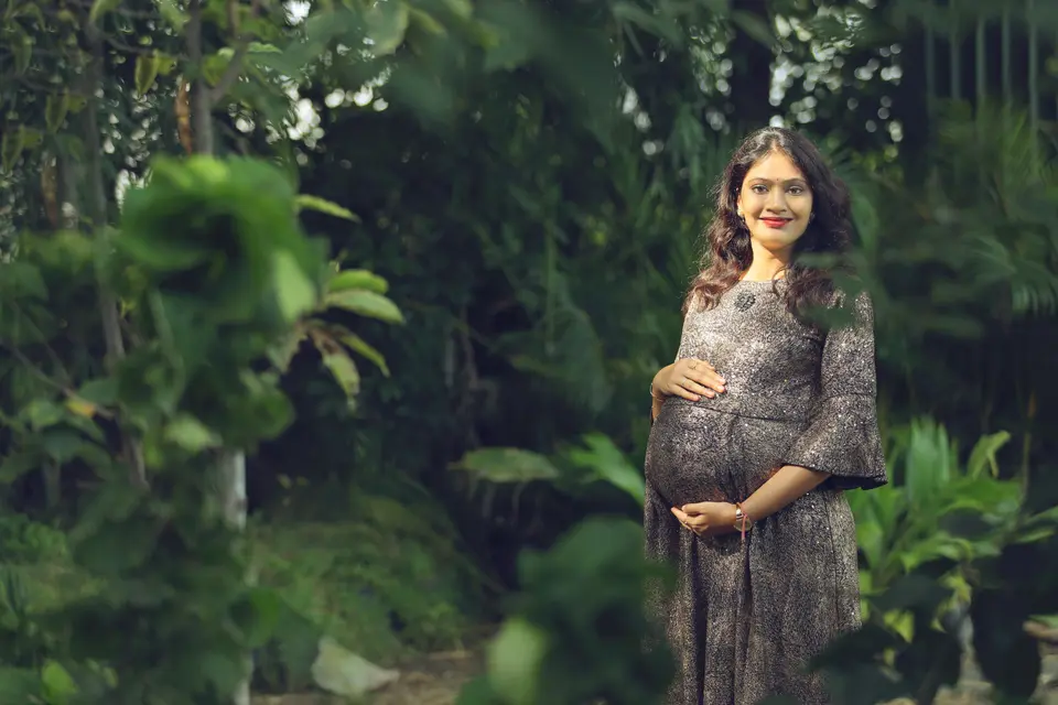 A smiling pregnant woman in a shimmering grey dress stands in a leafy garden with one hand resting on her belly