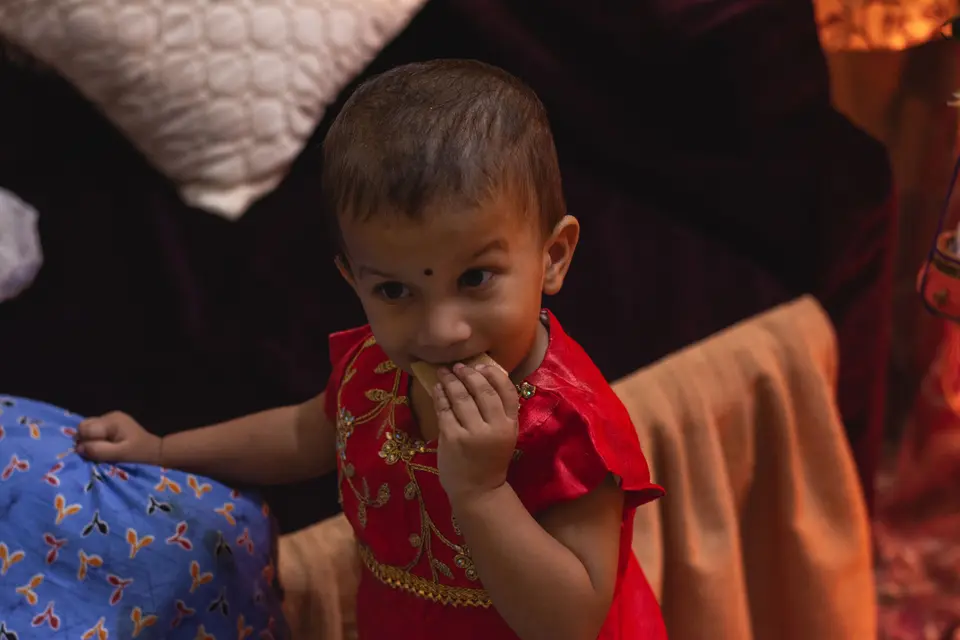 A South Asian toddler girl in a festive red embroidered dress eats a sweet snack by hand indoors.