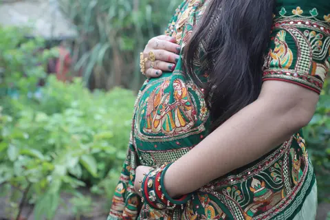 Close-up of a pregnant woman's belly in a green and gold embroidered outfit, her hand resting on top amid a green garden backdrop
