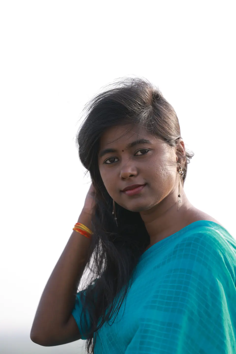 A South Asian woman in a teal sari stands in bright natural daylight, hand in her hair, with a calm and thoughtful expression.