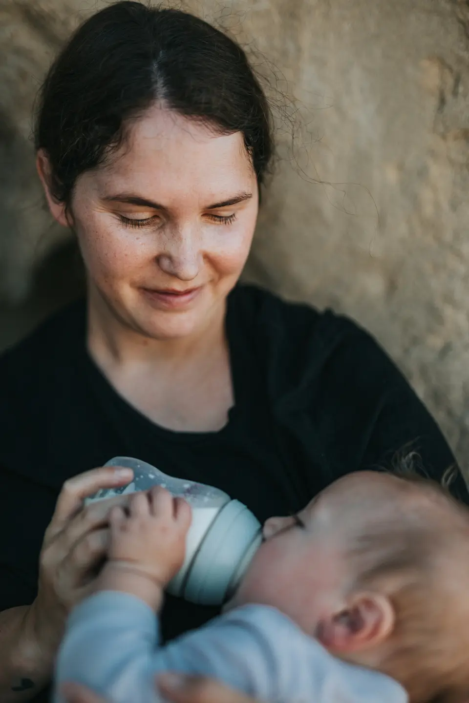 a mother smiles down at her baby while bottle-feeding, holding the baby securely upright against her chest with an unbranded bottle