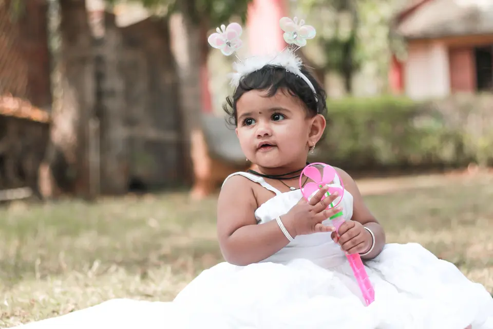 An Indian toddler girl in a white dress sits alone in a garden, absorbed in playing with a toy.