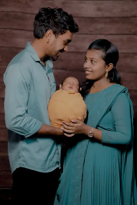 Parents in teal outfits smile at each other while holding their newborn, swaddled in mustard yellow, in an indoor studio portrait