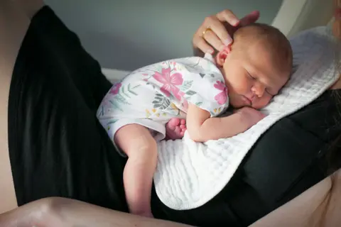 A mother cradles her sleeping newborn against her chest, one hand resting gently on the baby's head, seated indoors on a soft quilted wrap.