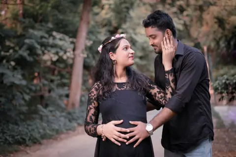 A pregnant woman in a black lace dress with a flower crown shares a tender moment with her partner outdoors, his hand on her cheek and hers on her belly
