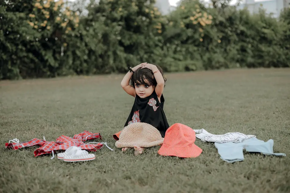 A South Asian toddler girl sits on the grass surrounded by clothes and hats, hands on her head as she decides what to wear.
