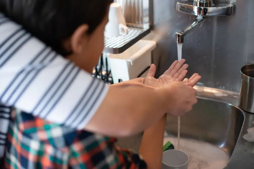 An adult helps a young child wash soapy hands at a kitchen sink under running water.