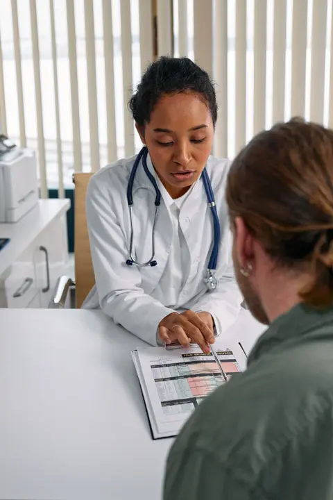 A female doctor reviews a printed chart across the desk with a seated patient during a consultation.