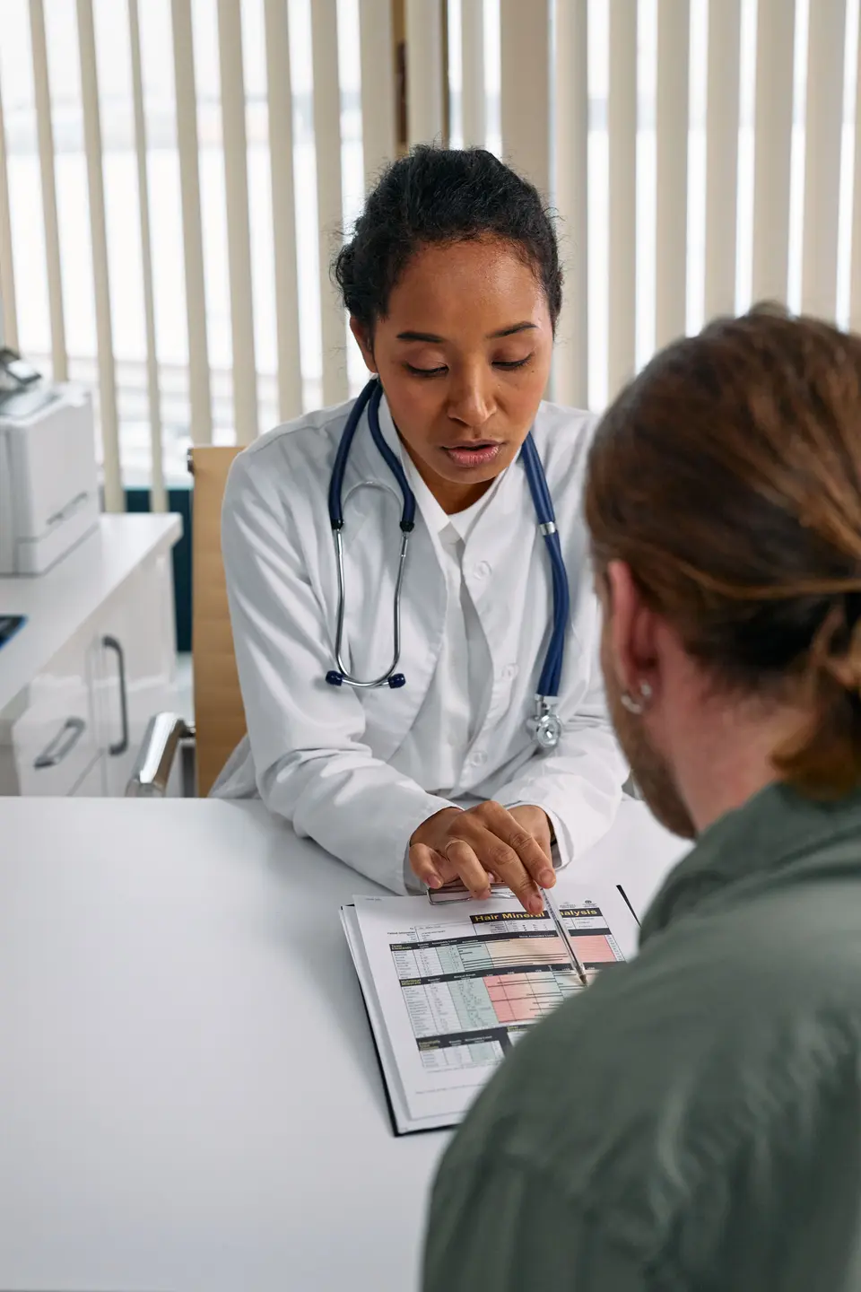 A female doctor reviews a printed chart across the desk with a seated patient during a consultation.