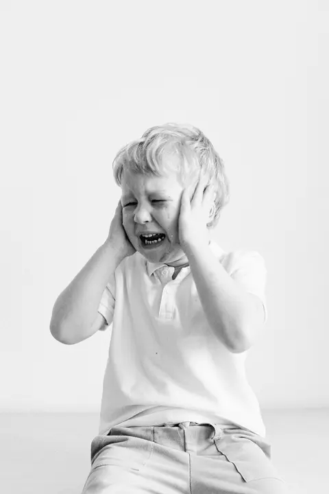 A black-and-white portrait of a young boy covering his ears while crying, showing an overwhelmed, dysregulated moment.