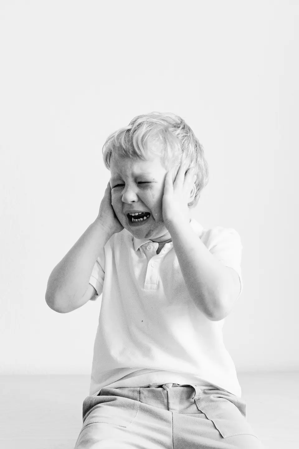 A black-and-white portrait of a young boy covering his ears while crying, showing an overwhelmed, dysregulated moment.
