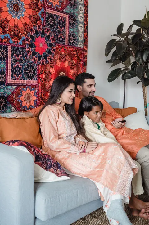 An Indian family of three sits close together on a sofa at home, the mother and father smiling warmly as their daughter watches TV between them.