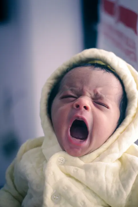 A South Asian baby in a cream hooded jacket cries with mouth wide open, close-up face portrait.