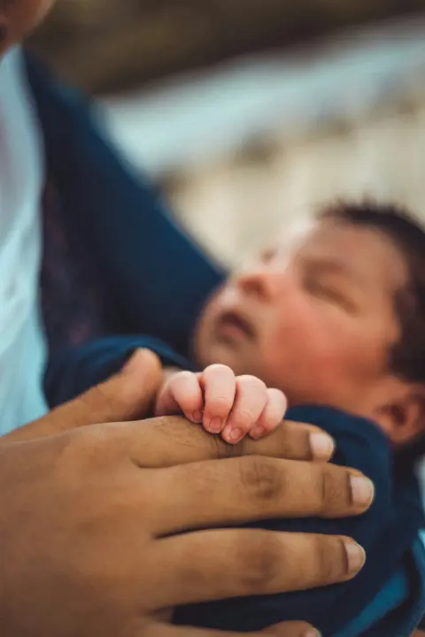 A father's hand cradles his newborn's tiny hand while the baby rests upright against his chest, close-up grayscale portrait.