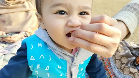 A South Asian toddler in a blue hooded top self-feeds a small piece of food held up by an adult's hand, outdoors.