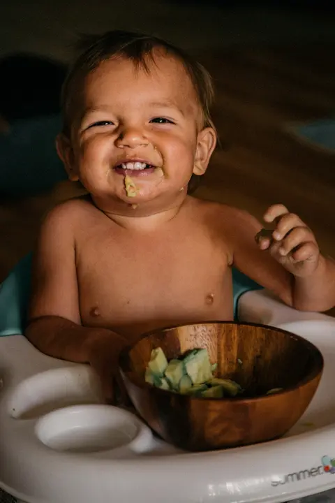 A baby sits in a feeding seat smiling with pureed green food on his face, holding a piece of food, with a wooden bowl of chopped vegetables in front of him.