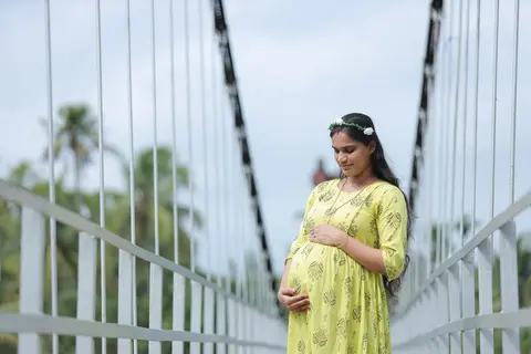 A visibly third-trimester pregnant South Asian woman in a yellow dress and flower crown stands on a footbridge, looking down and cradling her bare bump with both hands.