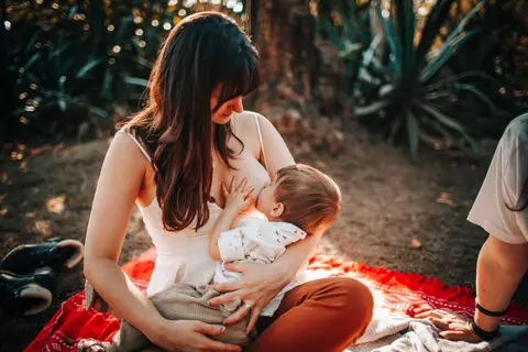 A mother breastfeeds her toddler-aged son outdoors in a forest clearing, warm natural sunlight, sitting on a red picnic blanket.