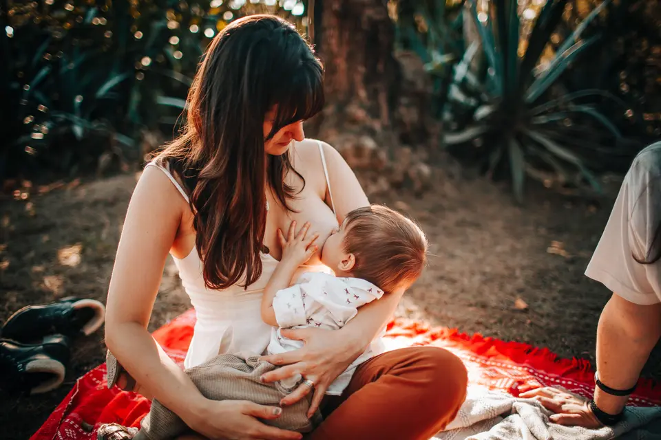 A mother breastfeeds her toddler-aged son outdoors in a forest clearing, warm natural sunlight, sitting on a red picnic blanket.