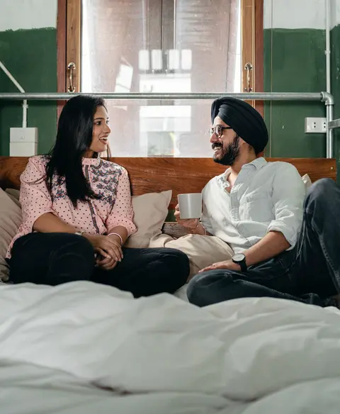 A Sikh man in a turban and a woman sit up in bed together, laughing and talking over coffee.