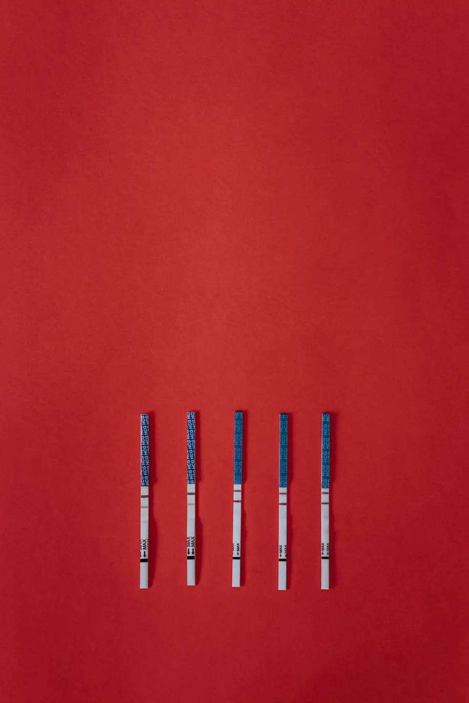 Five HCG/LH-style test strips with faint positive lines laid out flat against a bold red background.