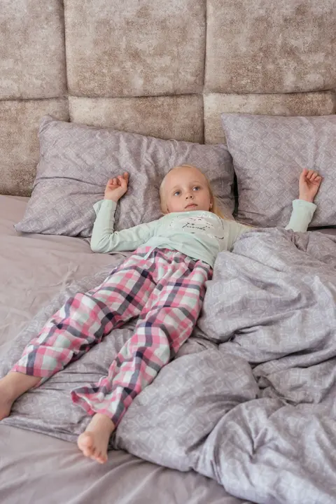 A young girl in checked pyjamas lies awake on a bed during the day, arms stretched out beside her pillow, in bright natural light.
