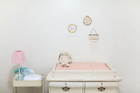 A plush bunny toy sits on a padded changing table in a softly decorated nursery, with a lamp and stacked diapers nearby