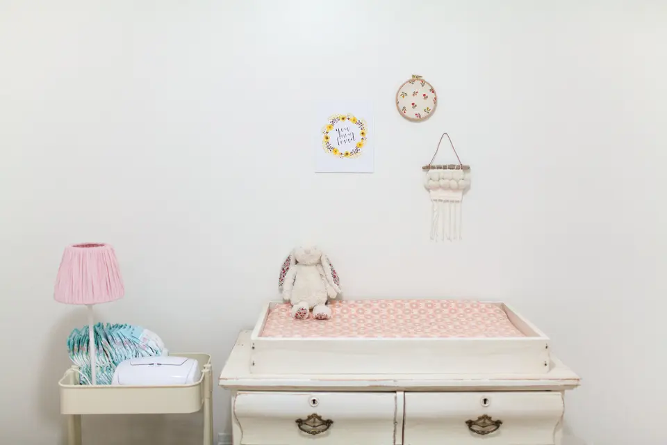 A plush bunny toy sits on a padded changing table in a softly decorated nursery, with a lamp and stacked diapers nearby