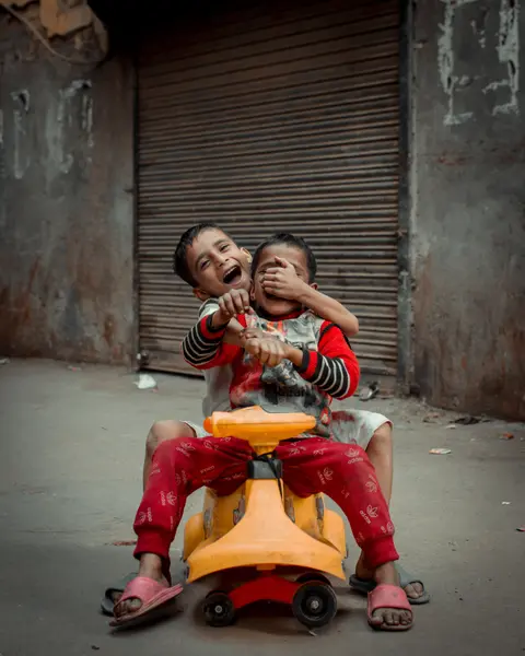 Two young Indian brothers laugh together while riding a shared plastic toy car on a street in New Delhi.